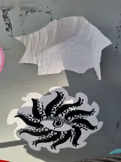 A black and white sticker featuring a striking design of an eye encircled by tentacle-like extensions, creating a sunburst or mandala-like effect. The style is reminiscent of occult or psychedelic art. The sticker is affixed to a gray surface, and a piece of another sticker is visible above it. 
