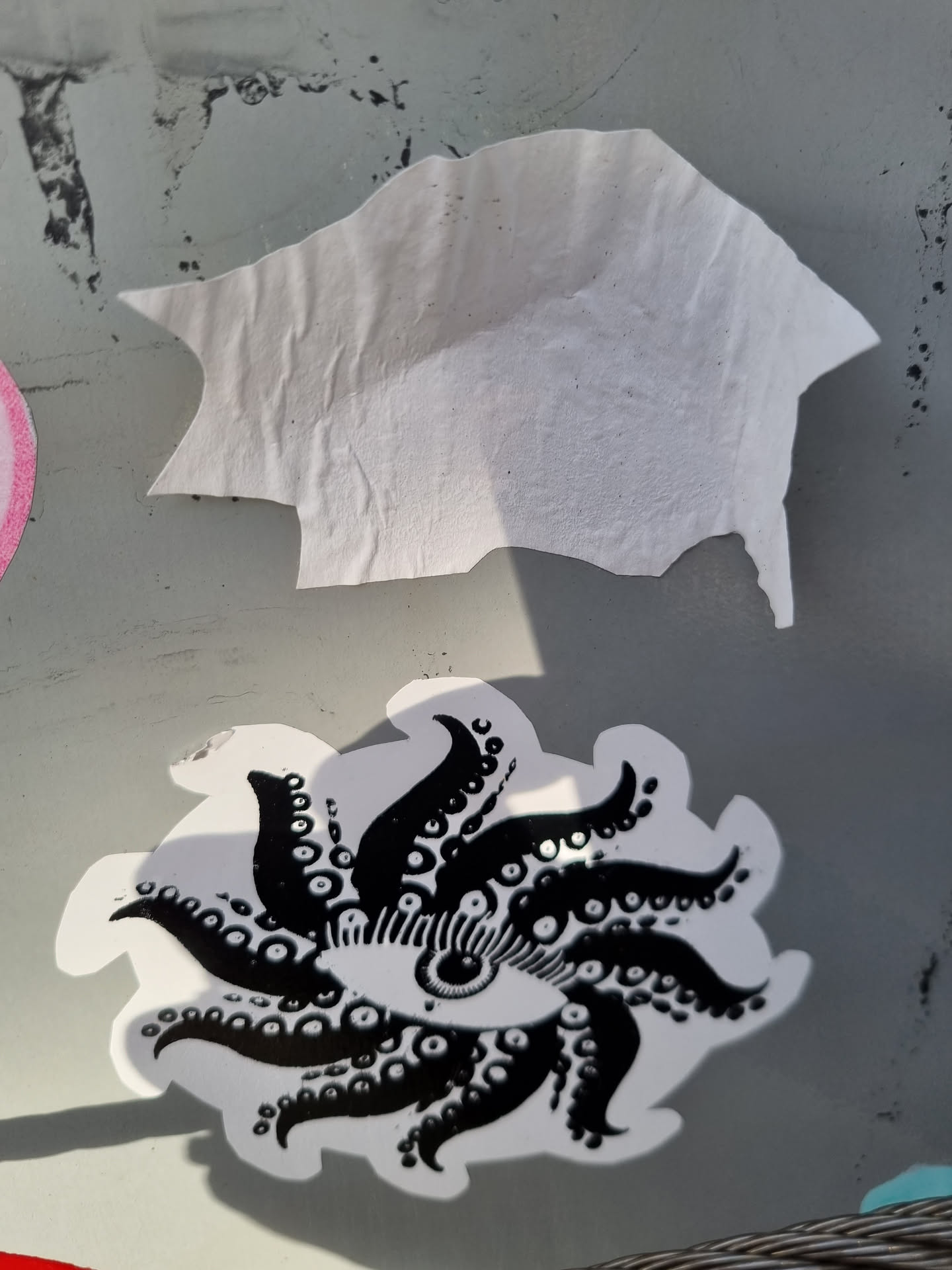 A black and white sticker featuring a striking design of an eye encircled by tentacle-like extensions, creating a sunburst or mandala-like effect. The style is reminiscent of occult or psychedelic art. The sticker is affixed to a gray surface, and a piece of another sticker is visible above it. 