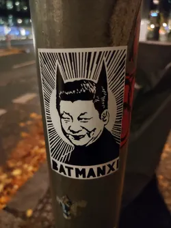 A sticker featuring a stylized image of Xi Jinping with bat ears, resembling Batman. The sticker is in black and white with the text "BATMANXI" at the bottom. 