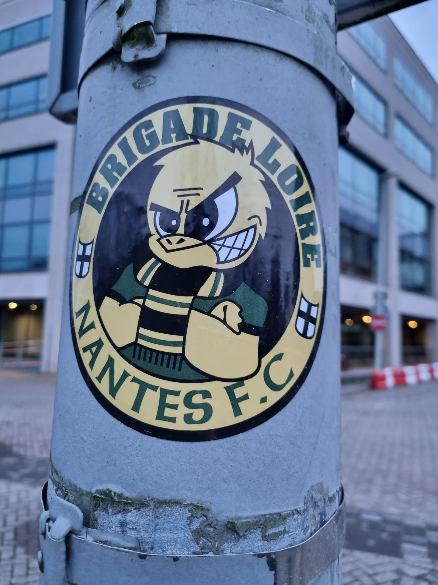 A circular sticker depicting the Nantes F.C. mascot, a cartoon duck with a stern expression, wearing a green and yellow scarf. The text 'BRIGADE LOIRE' is prominently displayed around the top of the circle, with 'NANTES F.C.' at the bottom. The sticker is affixed to a gray metal pole.
