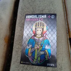 Holographic sticker featuring a stylized religious figure with flames, a crown, and X's over its eyes. The background is a metallic silver chain link fence.