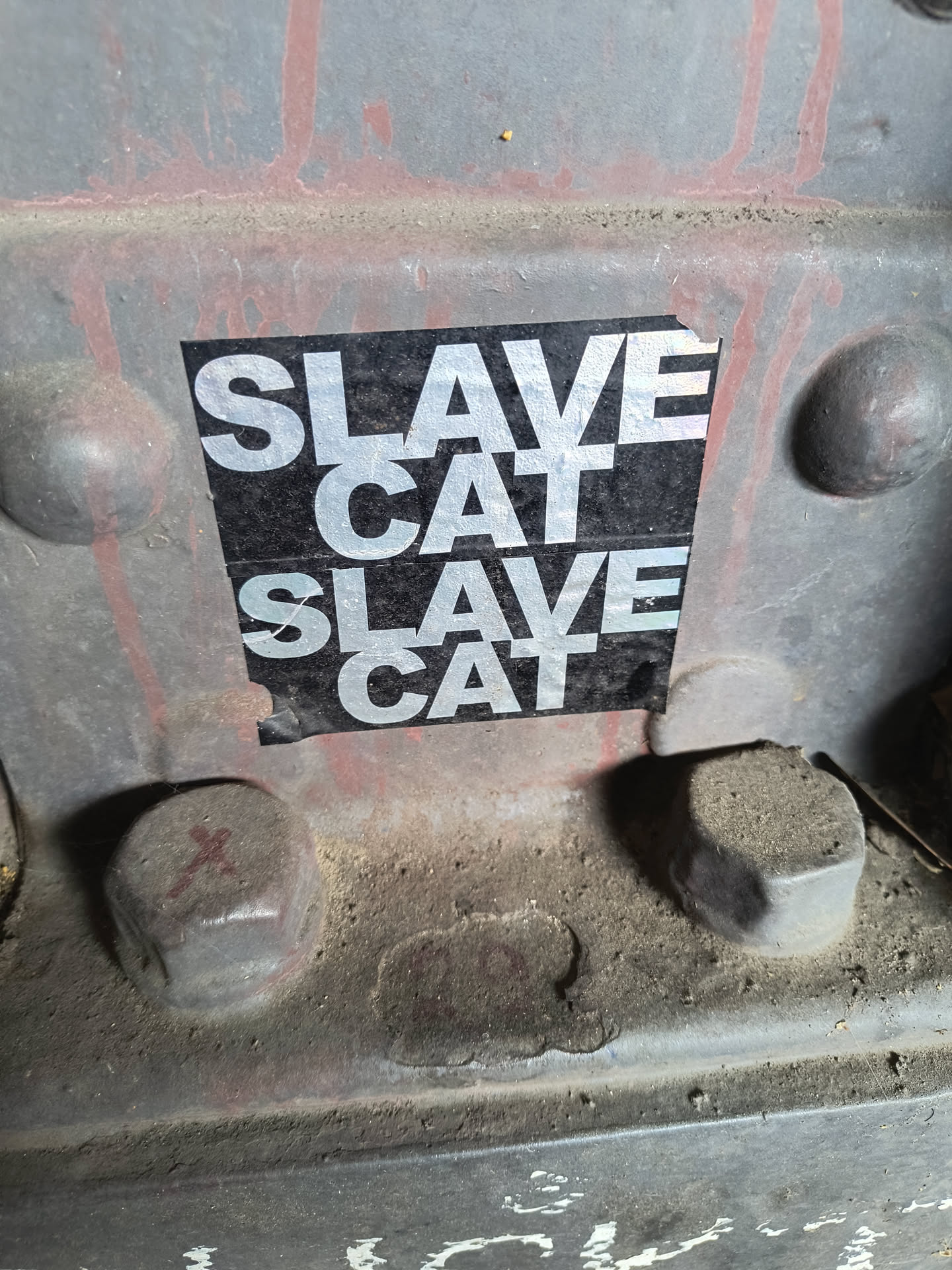 The sticker is rectangular with a black background and bold, silver capital letters. The text 'SLAVE CAT' is repeated twice, one above the other. The sticker shows some wear and appears to be slightly peeling at the bottom right corner. It is adhered to a rough, textured, gray metallic surface that has some reddish streaks and is dusty.