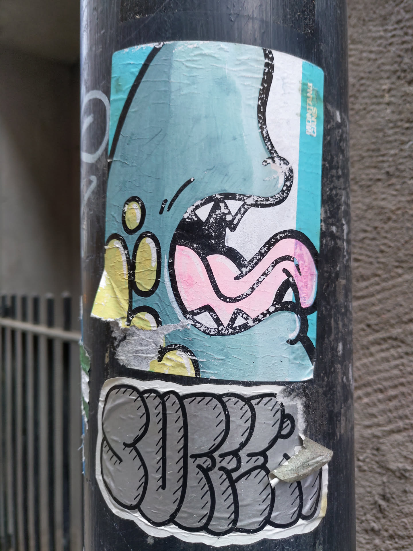 A sticker featuring a cartoon monster with a big mouth and tongue sticking out. The style is playful and colorful. Below the monster, the word "SURE" is written in stylized lettering. The sticker appears to be attached to a dark gray pole.