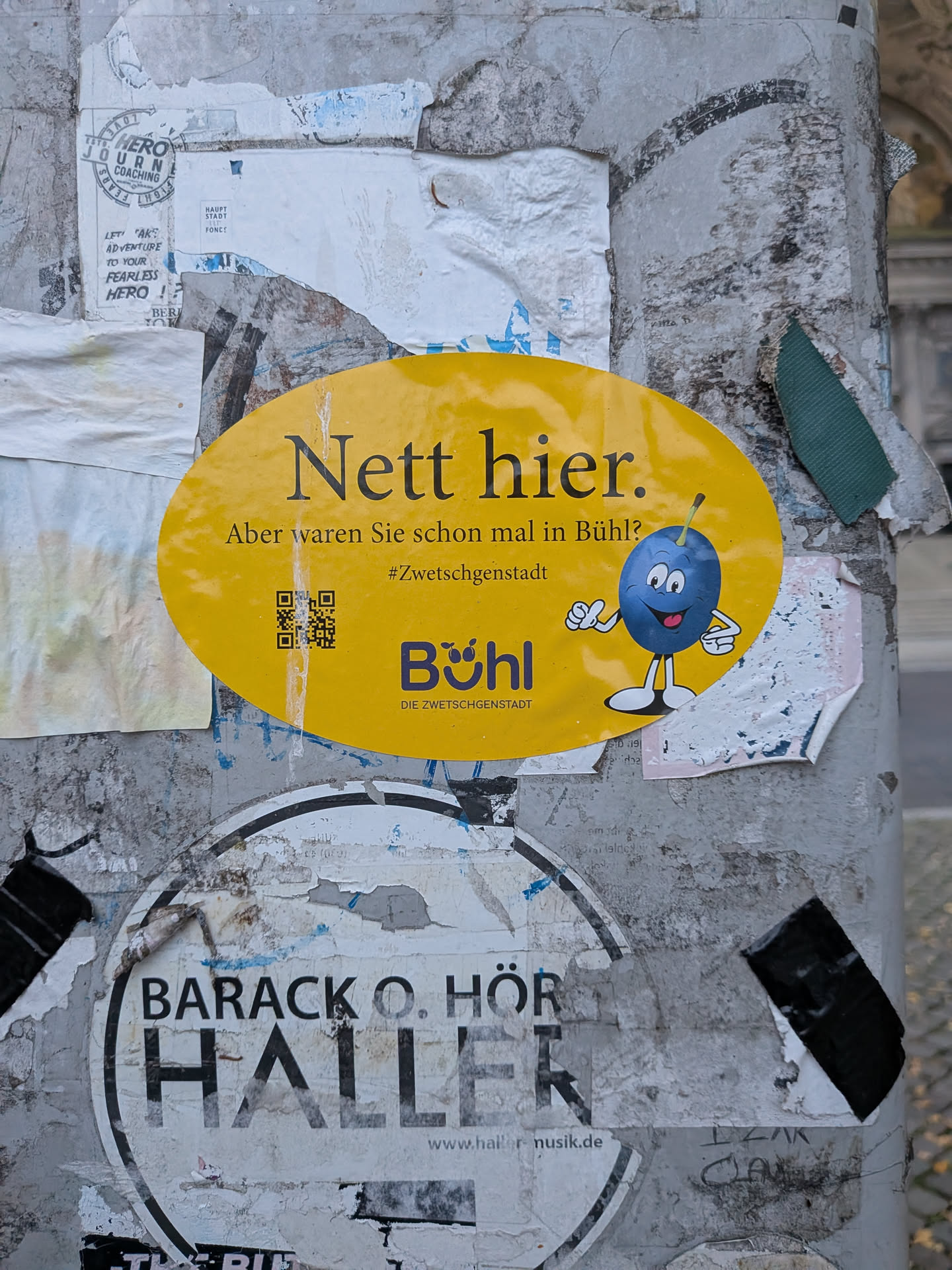 An oval-shaped yellow sticker mimicking the famous 'Nett hier. Aber waren Sie schon mal in Baden-Württemberg?' campaign. The sticker displays bold text reading 'Nett hier.' at the top, followed by 'Aber waren Sie schon mal in Bühl?' and the hashtag '#Zwetschgenstadt'. To the right is an anthropomorphic blue plum cartoon character with white gloves giving a thumbs up and white sneakers. Below the text is the city logo 'Bühl DIE ZWETSCHGENSTADT' and a small black QR code to the left.