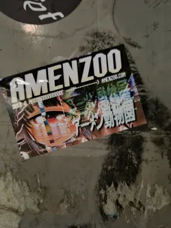 A sticker featuring the word 'AMENZOO' in bold white lettering, with a website address (amenzoo.com) and Japanese text below.  The sticker also displays vibrant anime artwork of a girl with a stylized digital glitch effect.