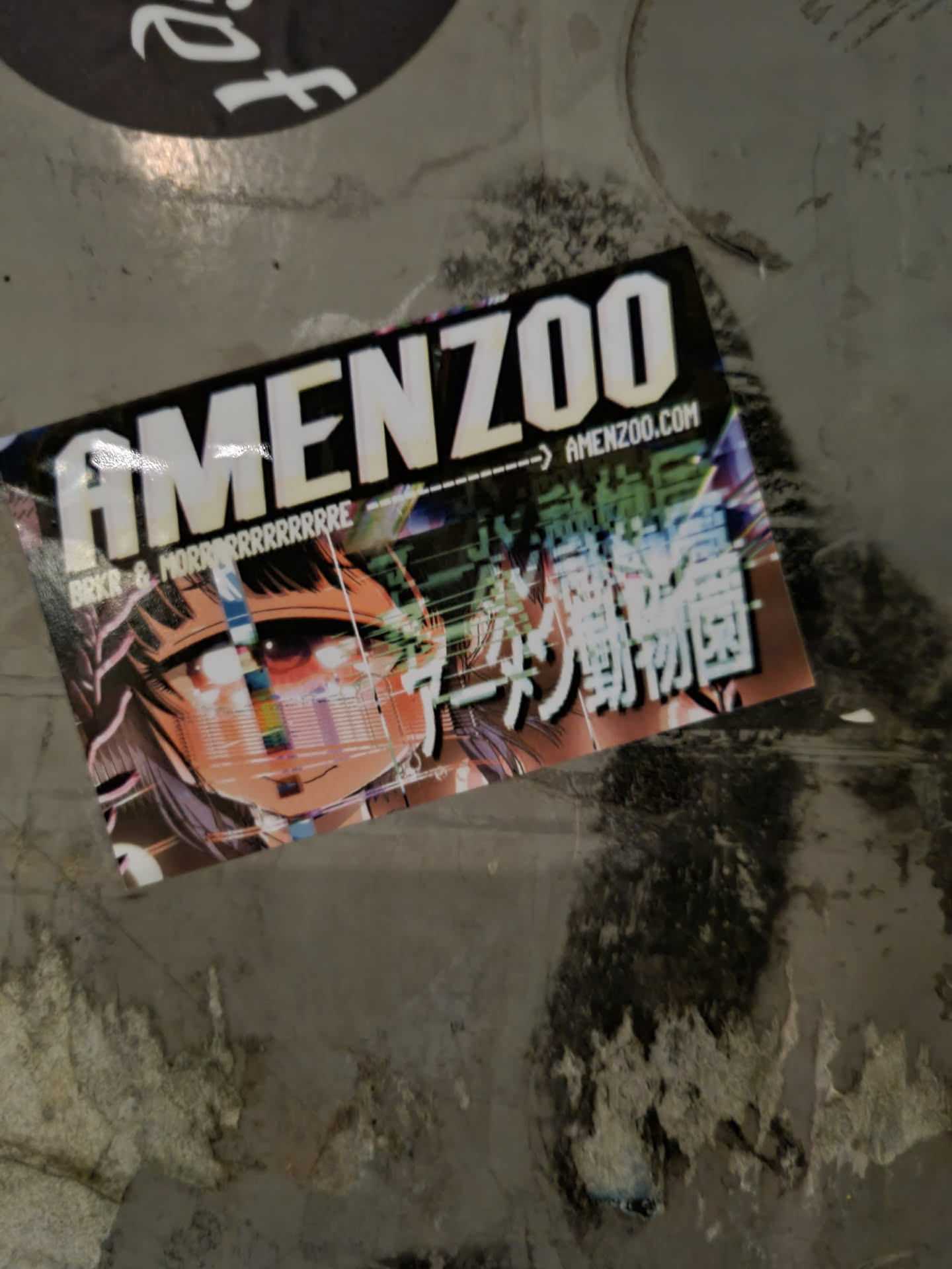A sticker featuring the word 'AMENZOO' in bold white lettering, with a website address (amenzoo.com) and Japanese text below.  The sticker also displays vibrant anime artwork of a girl with a stylized digital glitch effect.