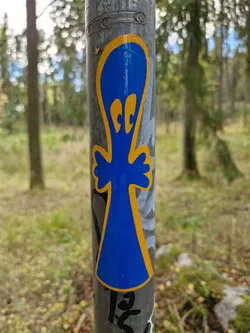 A blue ghost-like figure sticker with yellow outline, attached to a metal pole in a forest setting. The sticker has two small, round eyes in yellow.