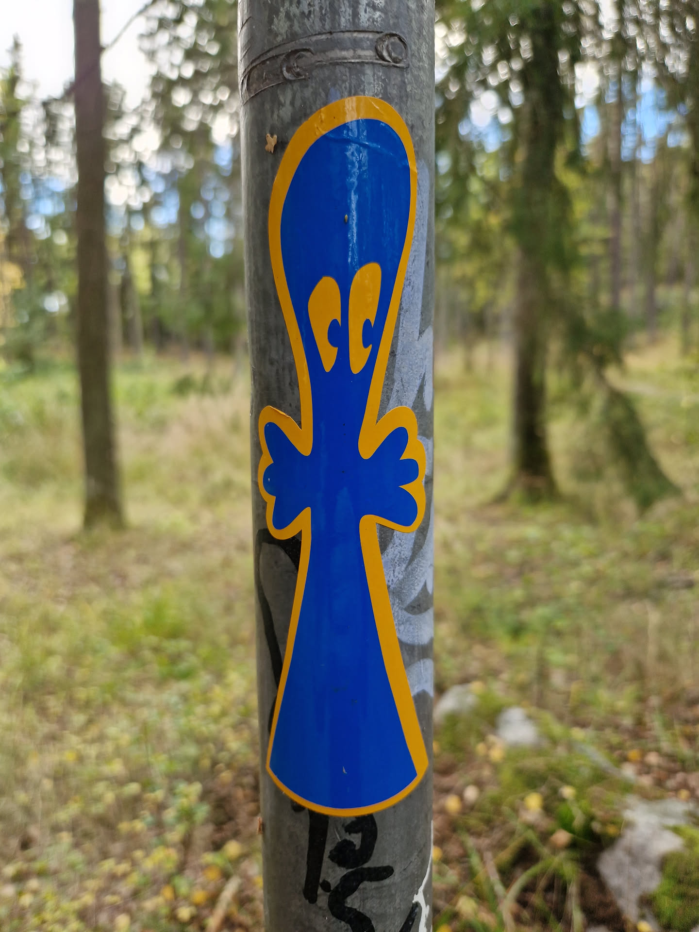 A blue ghost-like figure sticker with yellow outline, attached to a metal pole in a forest setting. The sticker has two small, round eyes in yellow.