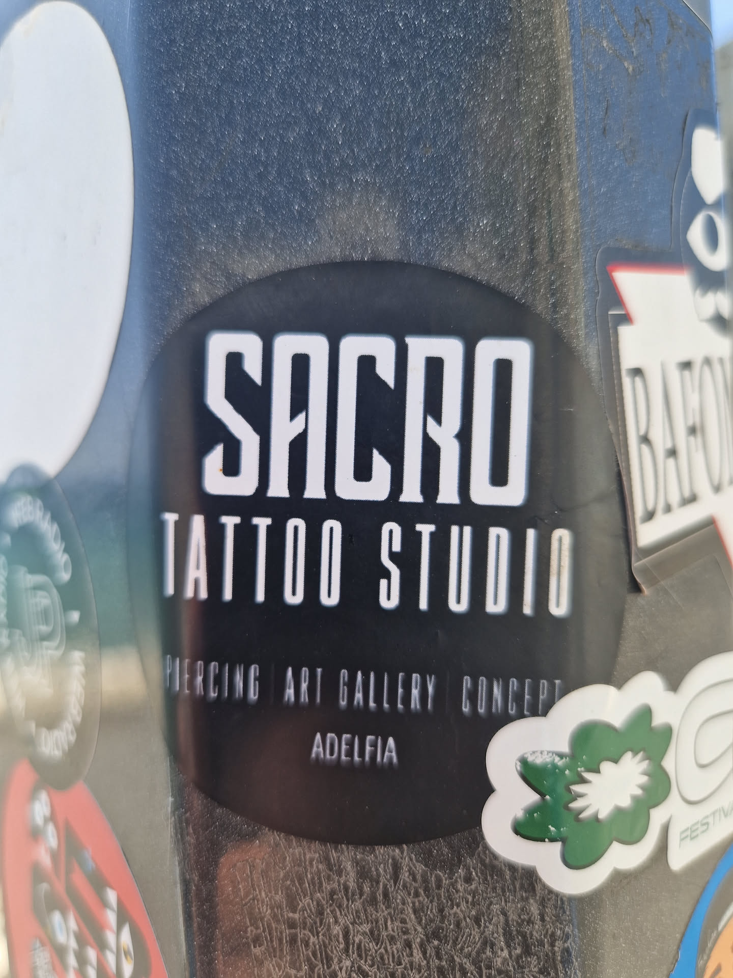Black and white sticker for Sacro Tattoo Studio in Adelfia, advertising their services: piercing, art gallery, and concept. The sticker has a round shape and features the studio's name and services in a minimalist design.