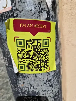 A yellow sticker with a QR code and the text "I'm an artist" on a dark pole. The sticker is slightly damaged.