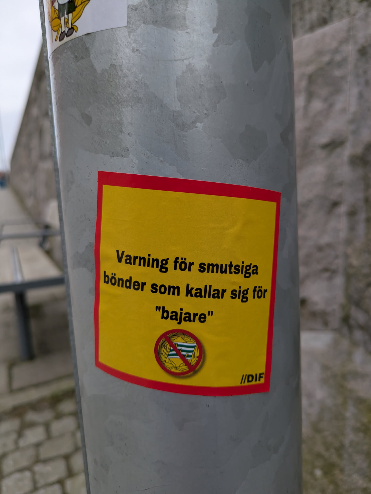 The sticker is rectangular with a yellow background and a red border. It warns in Swedish against "smutsiga bönder som kallar sig för 'bajare'", which translates to "dirty farmers who call themselves 'bajare'." Below the text is a symbol of the 'bajare' flag inside a crossed-out circle. In the bottom right corner, the text "//DIF" is visible.