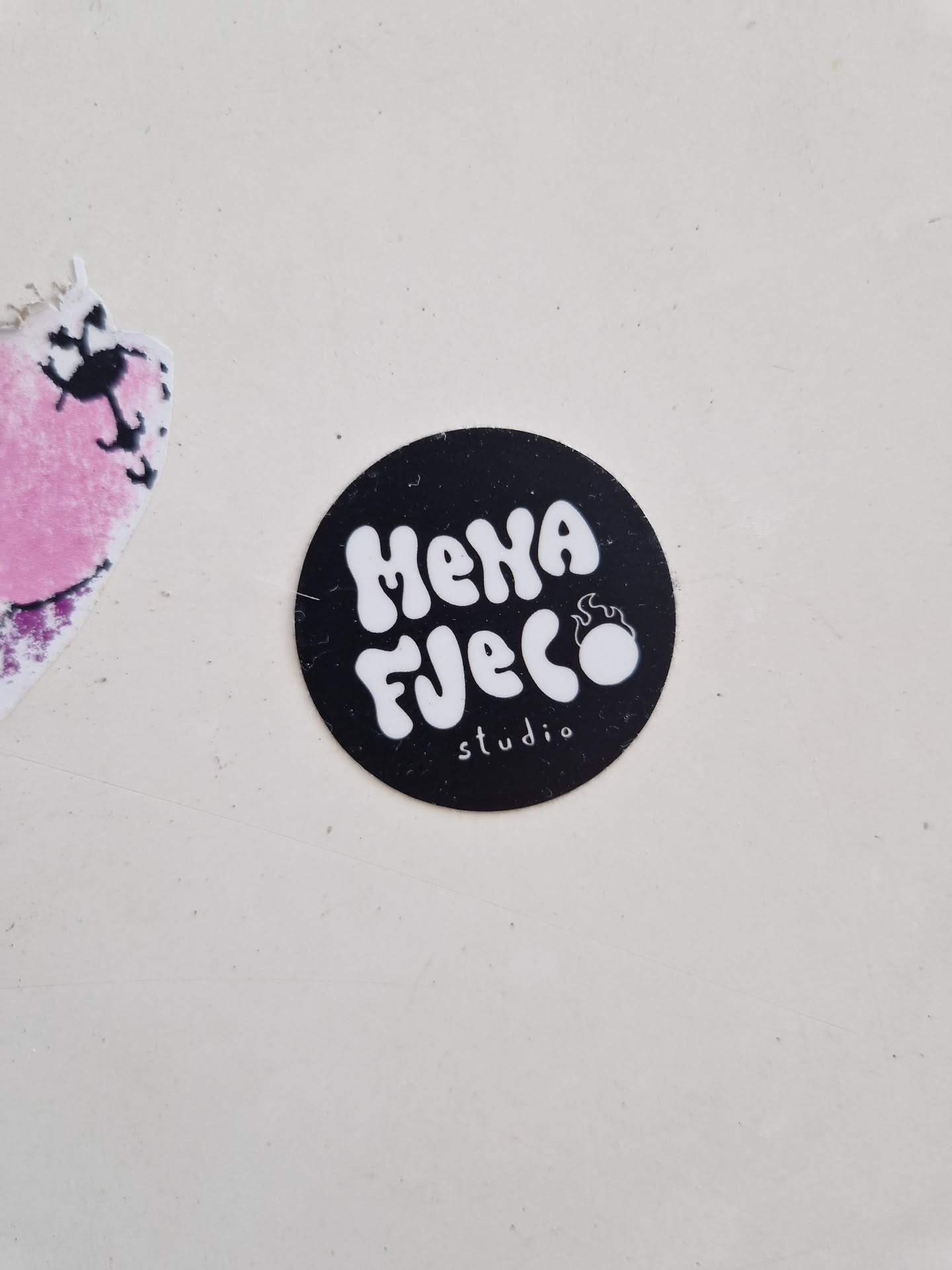 Close-up of a black and white circular sticker with the text "Meha Fuego studio" in a stylized font. The text is white and the background of the sticker is black. There is a small flame design next to the text.