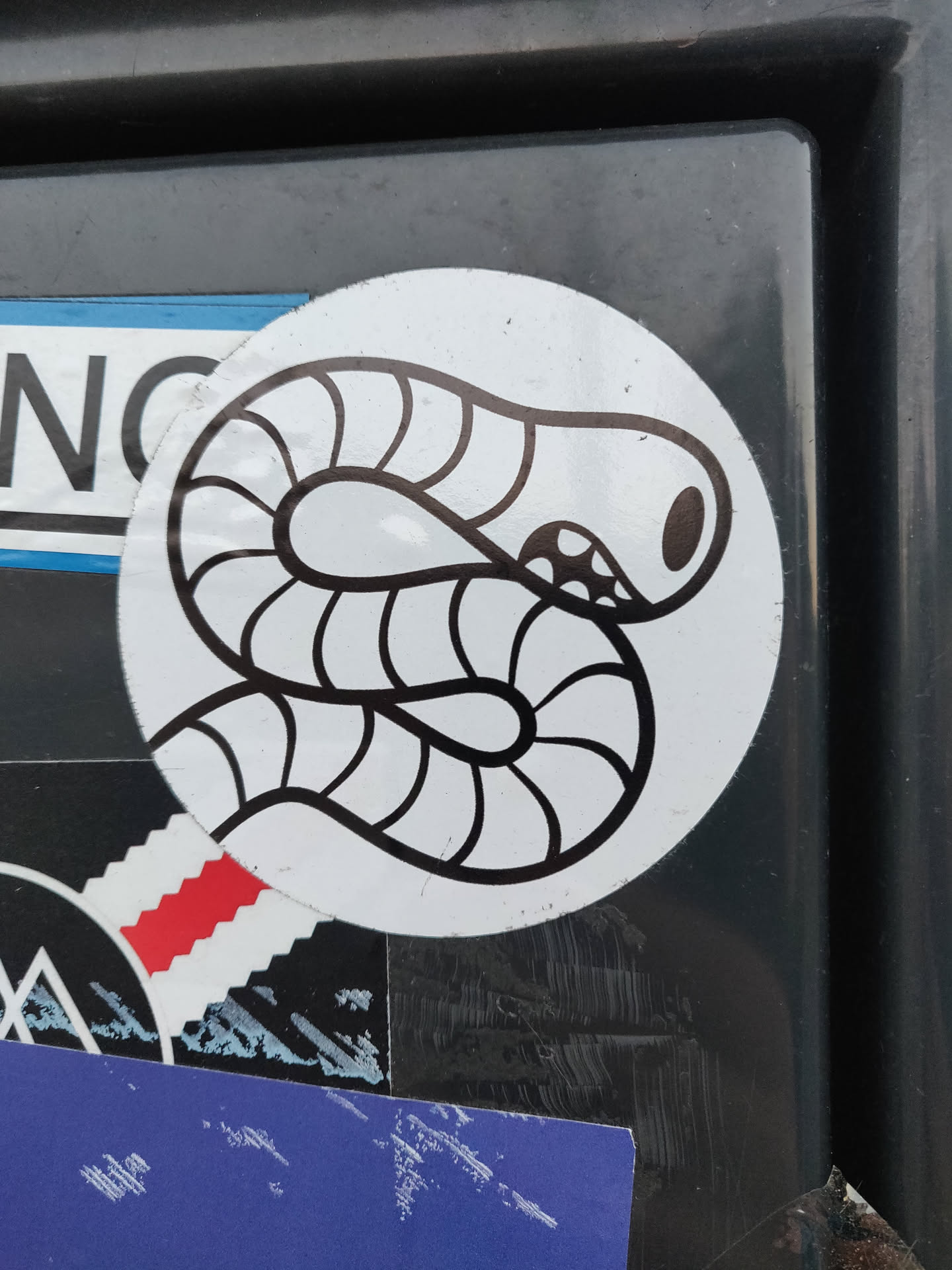A round sticker featuring a simple line drawing of a worm. The worm is depicted in black and white on a white background, with the worm's body segmented and curled around itself. The sticker appears to be placed on a dark gray or black surface, and other stickers are visible in the background. 