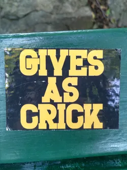 A sticker with the text "Gives as Crick" on a black background with yellow letters. The sticker is attached to a green surface.