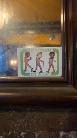 Street sticker A rectangular white sticker enclosed in a green rounded border, adhered to the bottom corner of a framed mirror. Inside the border are three humorous collage-style figures walking from left to right, each consisting of oversized heads pasted onto shortened bodies wearing shorts. The top banner reads 'MARTINS BARCRAWL' and the bottom banner reads 'KNÆ OG BRÆK' in green lettering.
