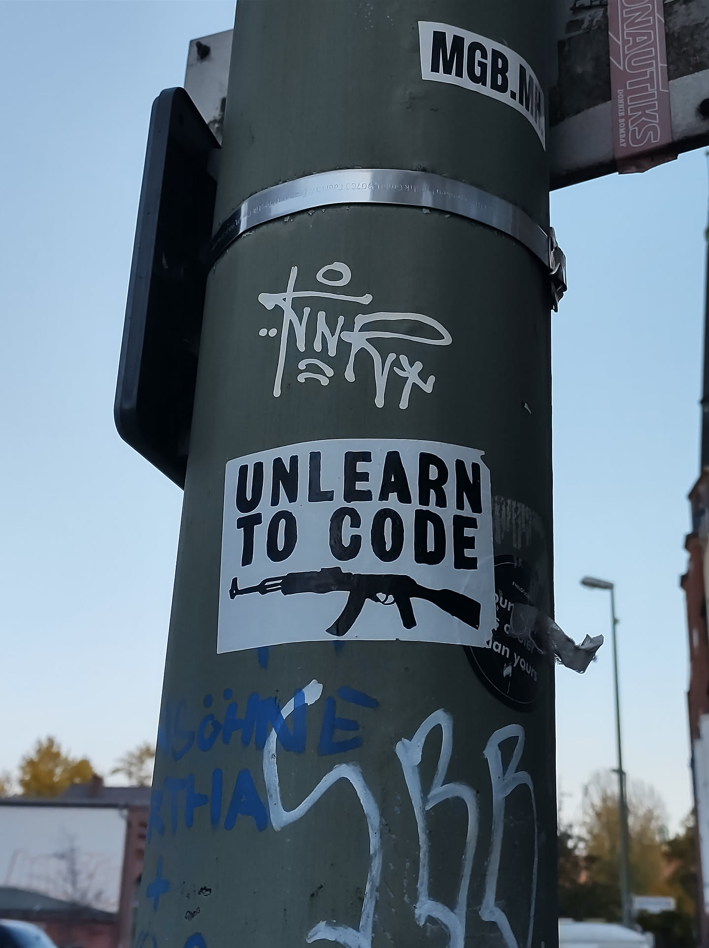 A sticker with the text "Unlearn to Code" and an image of an AK-47 rifle. The sticker is affixed to a pole with other graffiti and stickers.