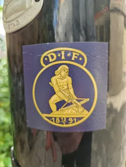 This sticker is rectangular and primarily dark purple, with a circular golden emblem in the center. The emblem features a detailed illustration of a man using a catapult, surrounded by text that includes "D.I.F." and "1812 91." The style evokes a historical or possibly military theme.
