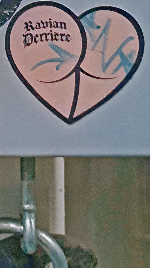 The sticker is shaped like a heart and is divided into two symmetrical sections resembling a pair of buttocks. The text 'Ravian Derriere' is written in a gothic-style font. There are also stylized arrows pointing towards the center of each 'cheek'.