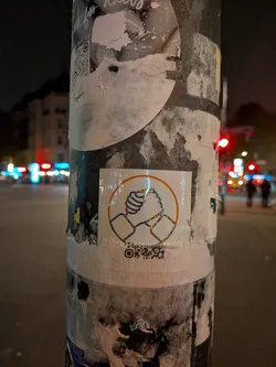 A square sticker affixed to a weathered post, featuring a circular motif in orange. Inside the circle, an outline of two hands firmly clasping each other in a gesture of unity or solidarity is rendered with sparkling, rainbow-holographic glitter lines. At the bottom of the sticker, an incomplete and partially cut-off QR code is printed.