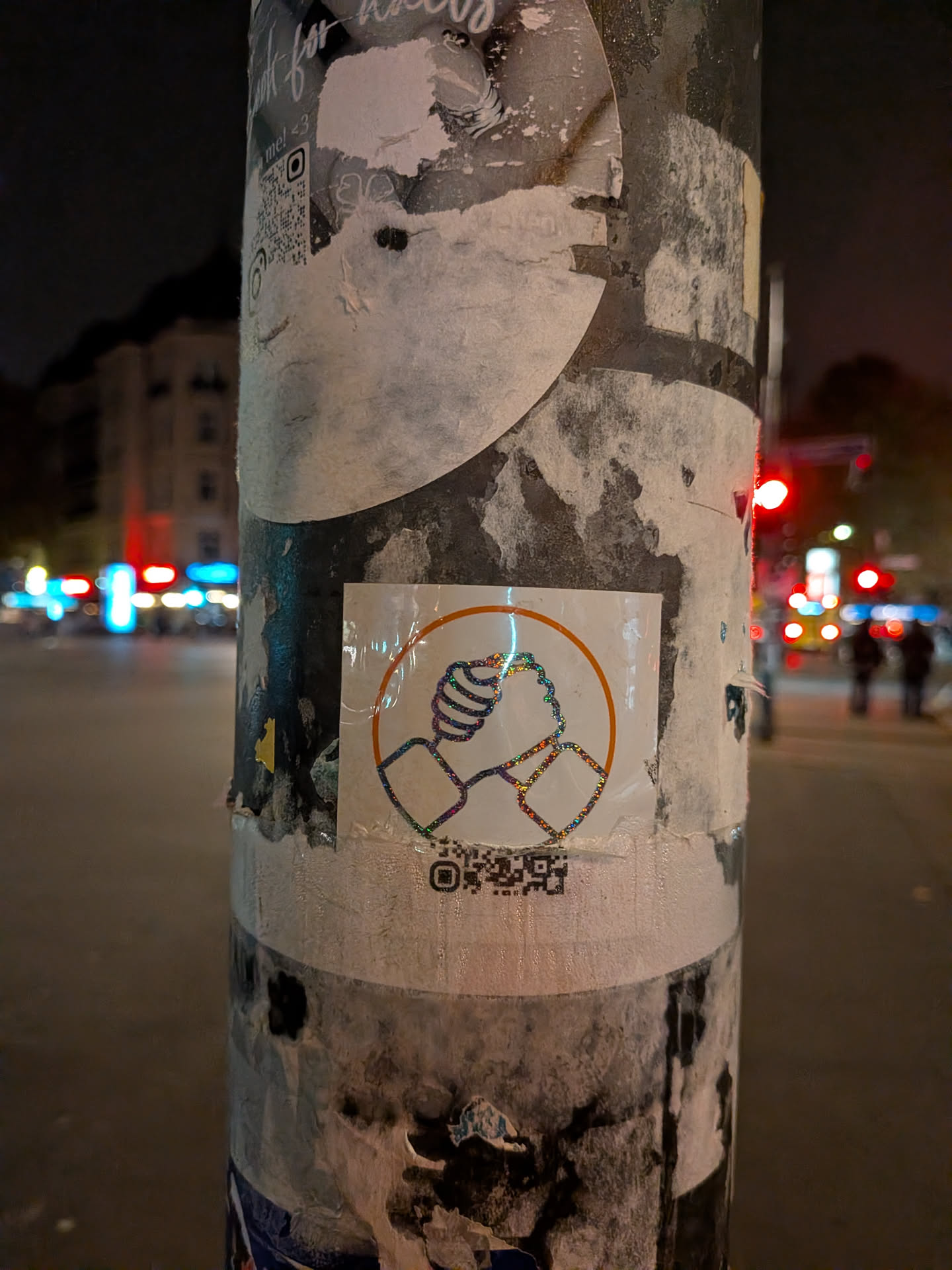 A square sticker affixed to a weathered post, featuring a circular motif in orange. Inside the circle, an outline of two hands firmly clasping each other in a gesture of unity or solidarity is rendered with sparkling, rainbow-holographic glitter lines. At the bottom of the sticker, an incomplete and partially cut-off QR code is printed.