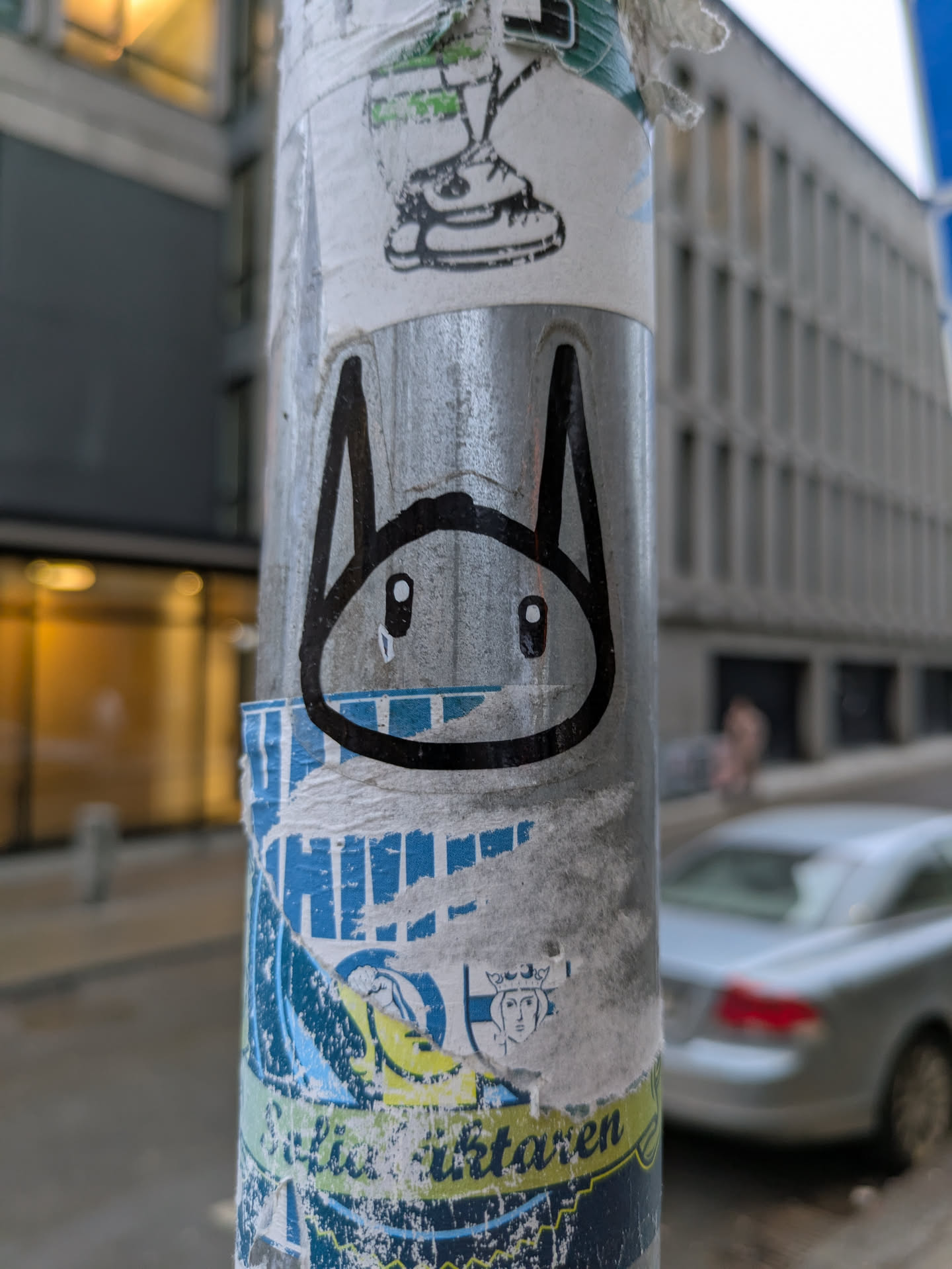 A sticker on a light pole features a black and white illustration of a bunny's face. The bunny has large, oval eyes and long, pointed ears. The style is simple and cartoonish. The sticker is placed among several other layers of older stickers.