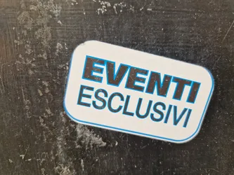 A sticker with the text "EVENTI ESCLUSIVI" (Exclusive Events) in Italian. The sticker is rectangular with rounded corners and has black text outlined in light blue on a white background. It's affixed to a dark, textured surface that appears to be slightly dirty or worn.