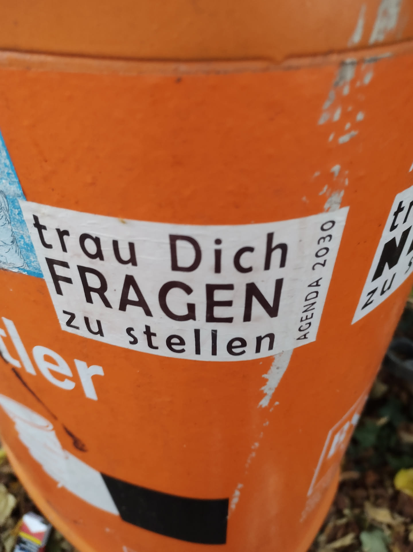 A sticker on an orange surface with the text "trau Dich FRAGEN zu stellen" (Dare to ask questions) in German.  There is also a small Agenda 2030 reference visible on the sticker.