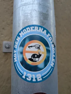 Street sticker A circular sticker affixed to a round metal pipe. The outer border features light blue concentric bands with bold white capital letters reading 'MOT DEN MODERNA FOTBOLLEN' around the top edge and the number '1312' at the bottom. Inside the ring, set against a dark teal background, is a silhouette of a riot police officer wearing a helmet emblazoned with the Swedish police emblem, raising a baton inside a yellow-to-orange circular emblem.