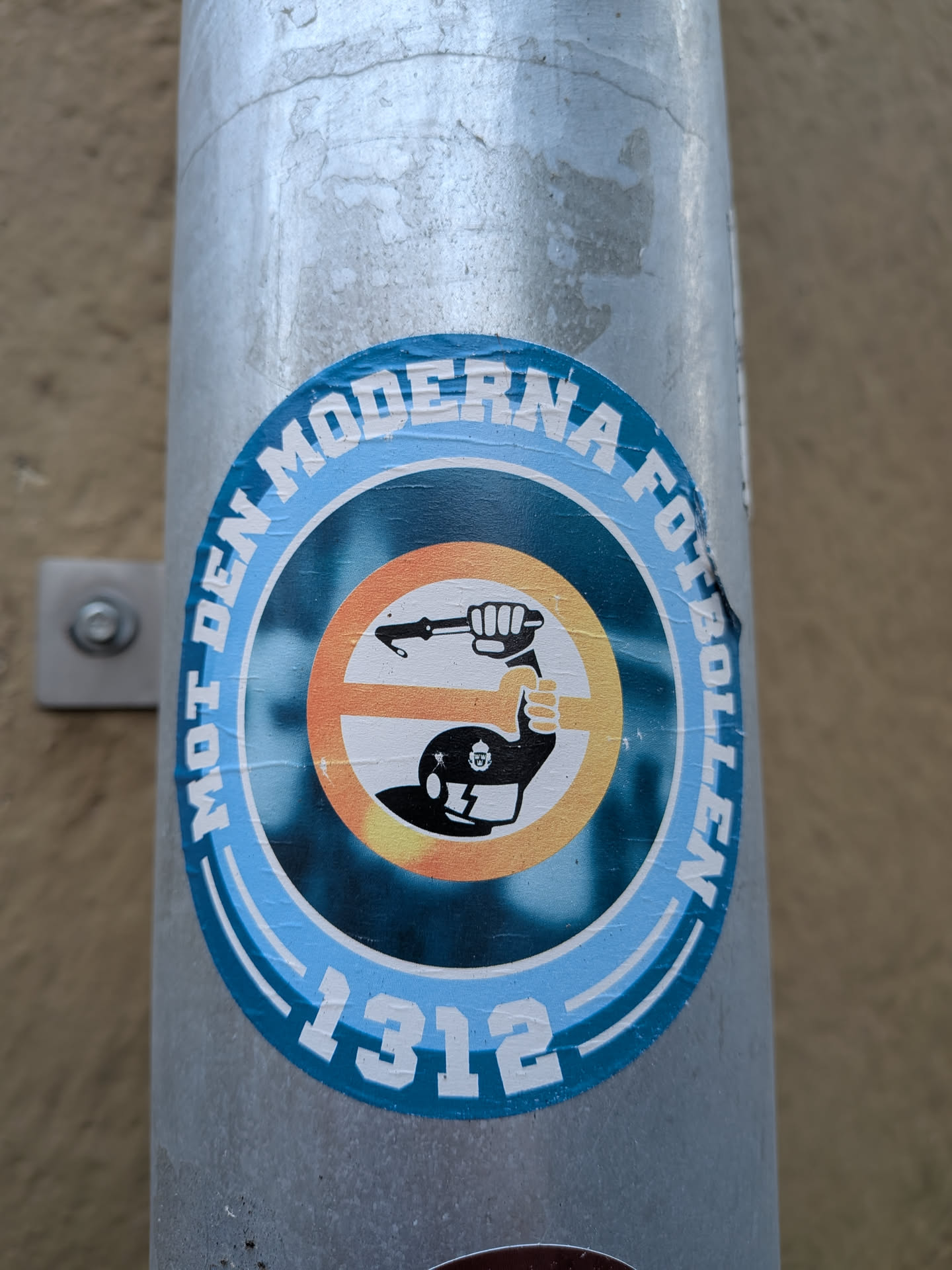 A circular sticker affixed to a round metal pipe. The outer border features light blue concentric bands with bold white capital letters reading 'MOT DEN MODERNA FOTBOLLEN' around the top edge and the number '1312' at the bottom. Inside the ring, set against a dark teal background, is a silhouette of a riot police officer wearing a helmet emblazoned with the Swedish police emblem, raising a baton inside a yellow-to-orange circular emblem.