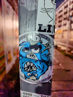 A sticker featuring a cartoon-like blue creature with an angry expression. The creature seems to have a combination of feline and monstrous features. The sticker is placed on a grey pole, with other stickers visible around it.