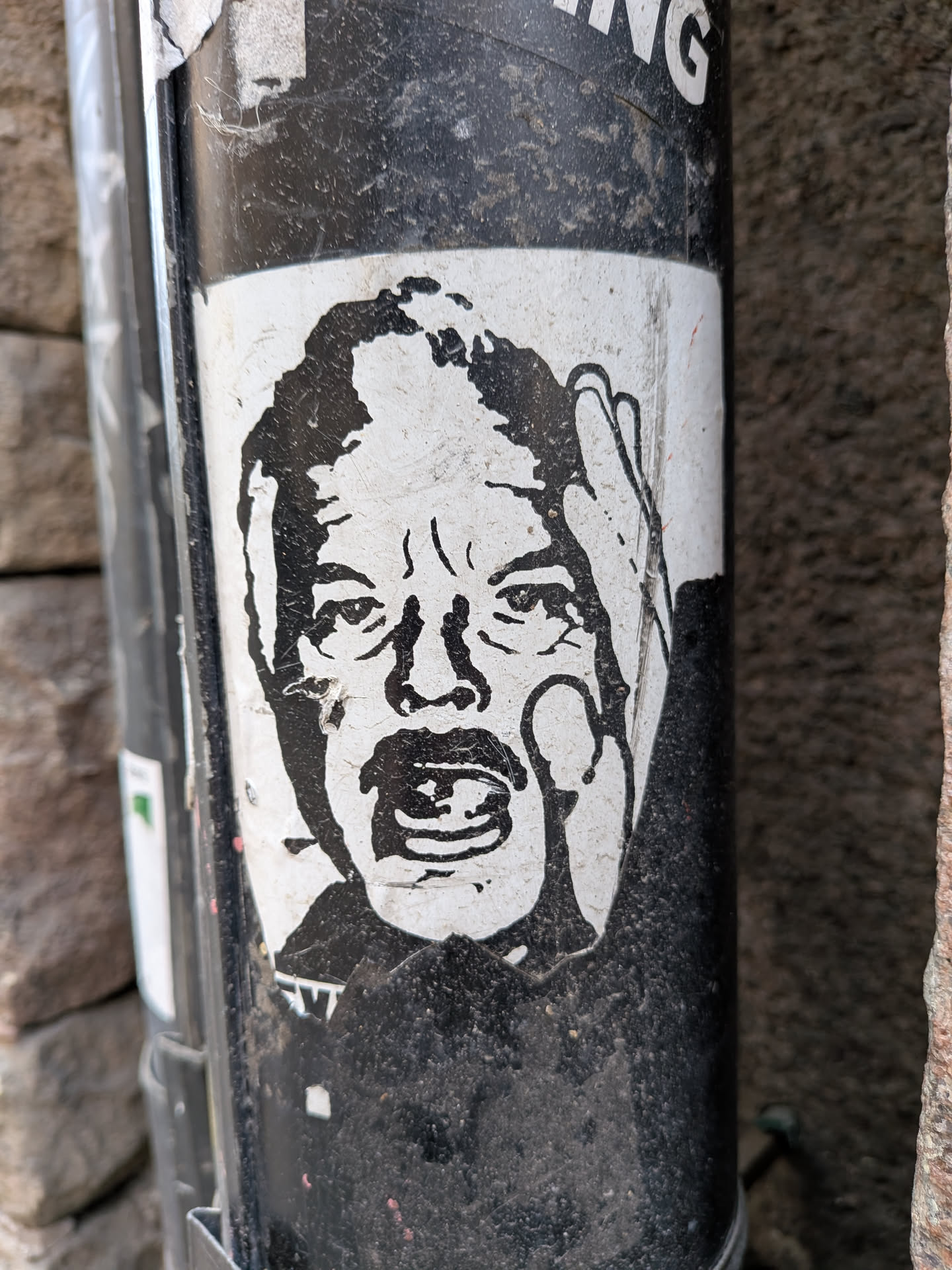 The sticker features a stark, black-and-white graphic of a man's head and shoulders. His face is contorted in an expression of intense shock or a scream, with his mouth wide open and eyes wide. One hand is raised to the side of his head, fingers spread, emphasizing the gesture of despair or surprise. The artwork has a rough, almost woodcut or stencil-like aesthetic, rendered in solid black shapes against a white background. The sticker itself shows signs of wear, with slightly peeling edges and some dirt accumulation, adhering to a textured, grimy black pole.