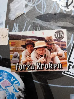 Street sticker A rectangular photographic sticker featuring three men crouching down and pointing intensely toward the viewer. They are dressed in khaki safari/fisherman style outfits with distinct hats. The background shows the local shopping center 'Krokensenteret' in Tromsø, Norway. In the upper right corner sits a black laurel wreath logo containing the letters 'TIL' (Tromsø Idrettslag). The text 'Forza Kroken' is displayed prominently across the bottom in a bold black Gothic font with a white outline.