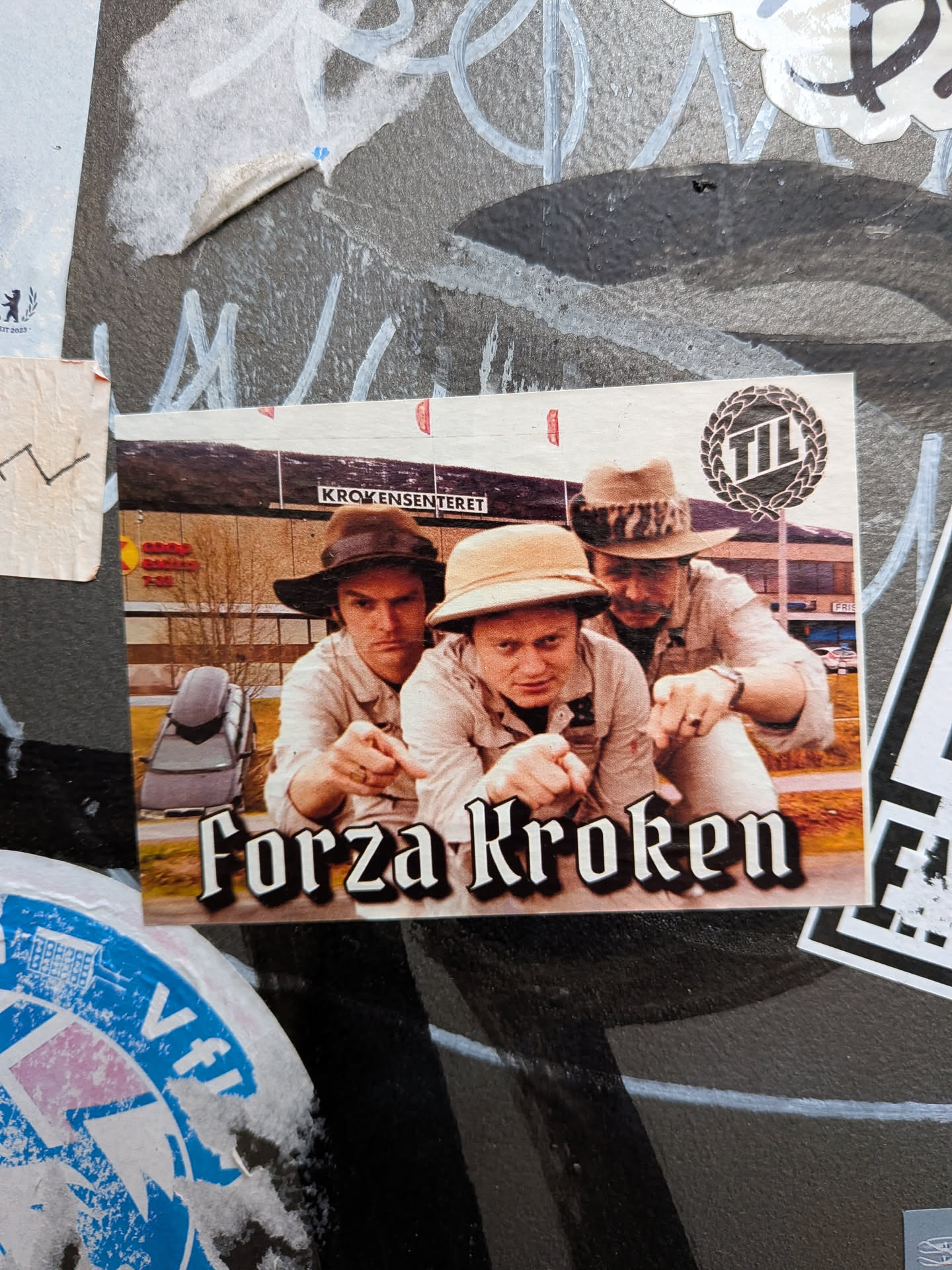 A rectangular photographic sticker featuring three men crouching down and pointing intensely toward the viewer. They are dressed in khaki safari/fisherman style outfits with distinct hats. The background shows the local shopping center 'Krokensenteret' in Tromsø, Norway. In the upper right corner sits a black laurel wreath logo containing the letters 'TIL' (Tromsø Idrettslag). The text 'Forza Kroken' is displayed prominently across the bottom in a bold black Gothic font with a white outline.