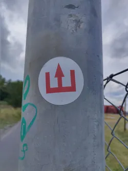 A round white sticker with a red symbol resembling an upward arrow between two vertical bars affixed to a grey pole. The symbol is reminiscent of a simplified castle or gate.