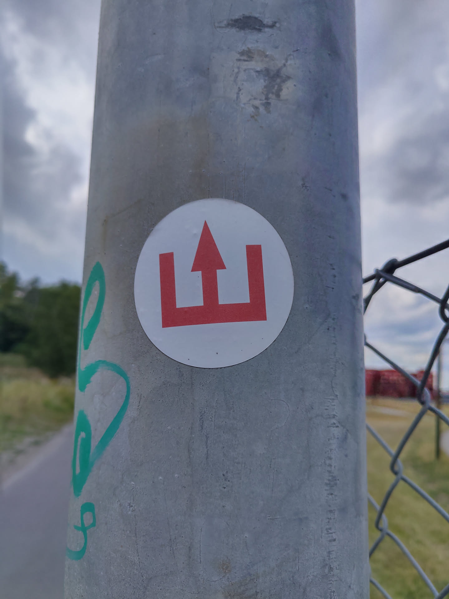 A round white sticker with a red symbol resembling an upward arrow between two vertical bars affixed to a grey pole. The symbol is reminiscent of a simplified castle or gate.