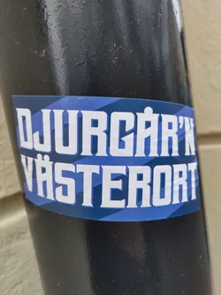 A sticker with the text "DJURGÅRDEN VÄSTERORT" in white bold letters on a blue and dark blue striped background. The sticker is attached to a black pole.