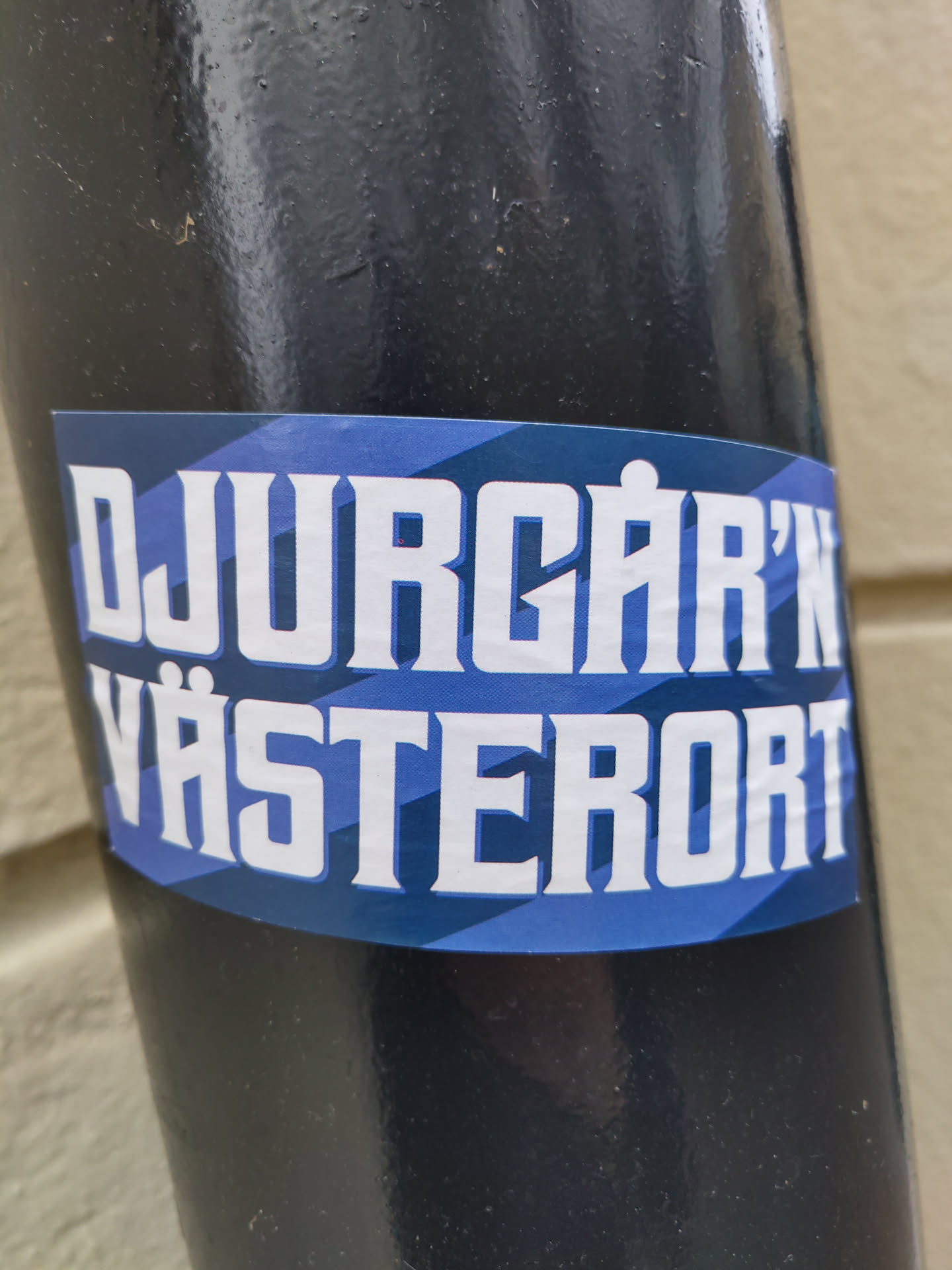 A sticker with the text "DJURGÅRDEN VÄSTERORT" in white bold letters on a blue and dark blue striped background. The sticker is attached to a black pole.