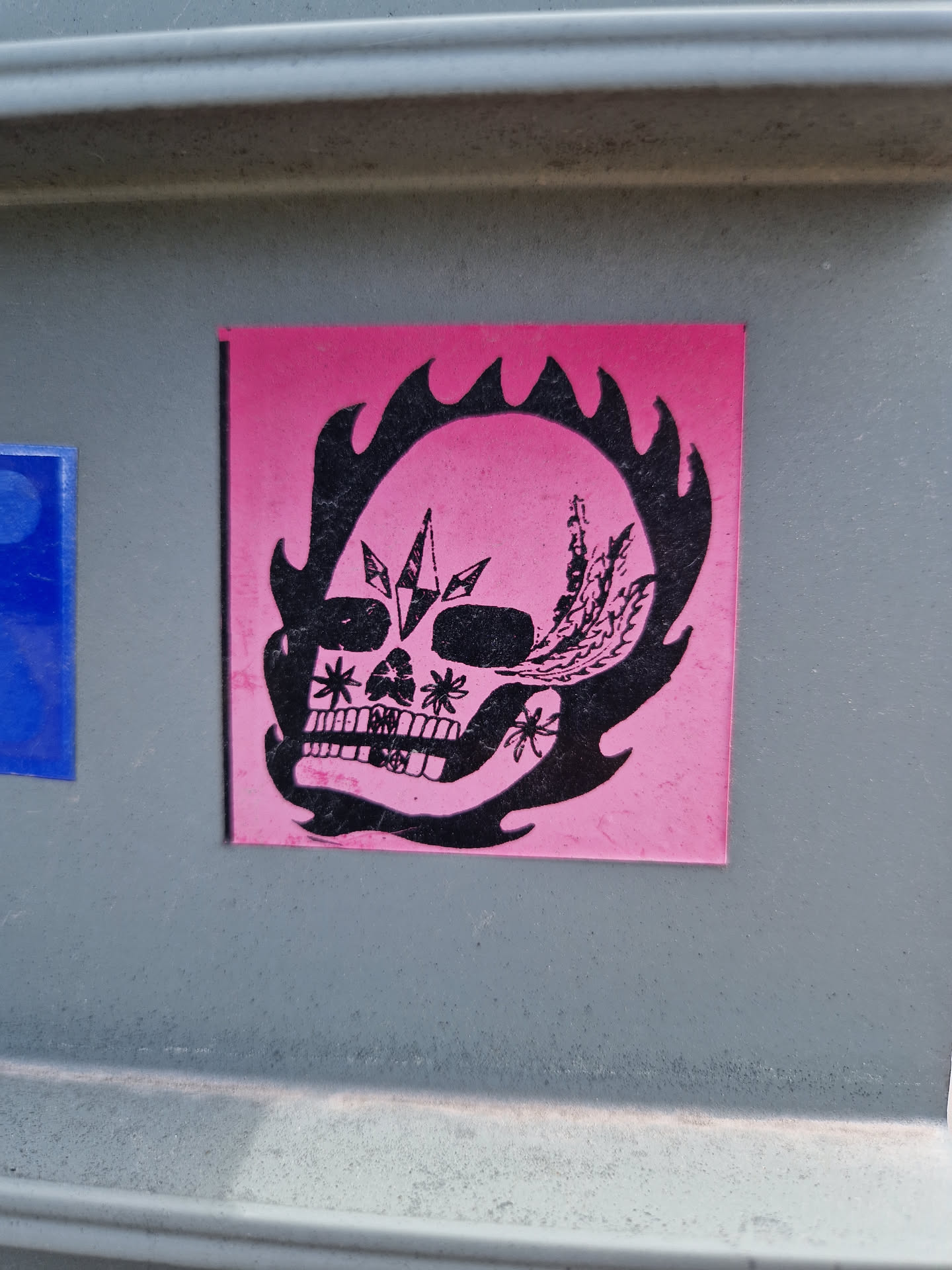A pink sticker with a black illustration of a skull engulfed in flames. The skull is adorned with crystals and floral details. 