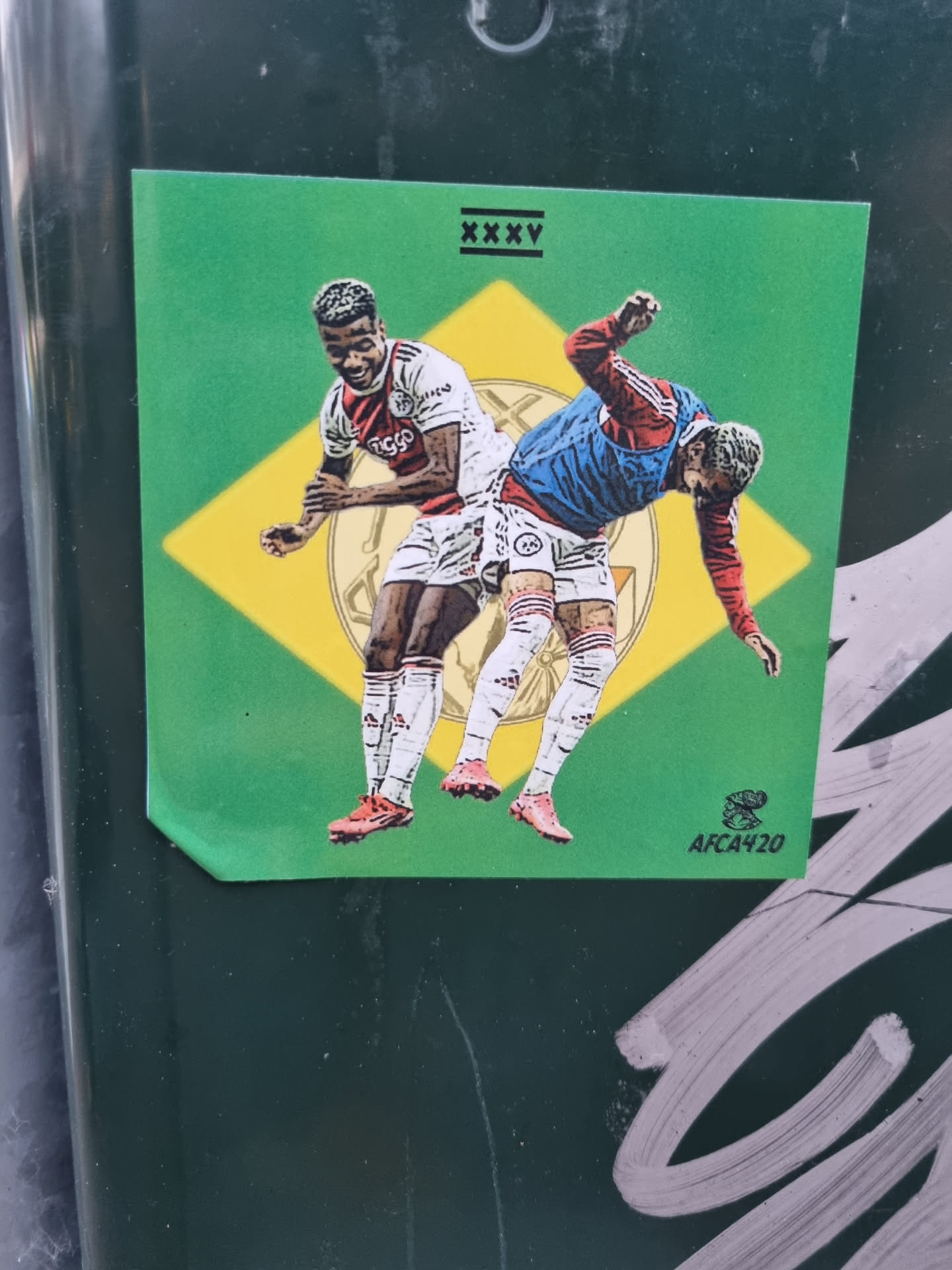 A sticker depicting a football match scene between Antony and Lisandro Martínez. The illustration style is reminiscent of trading cards or limited-edition prints. The sticker has a green background with yellow accents, and a small logo 'AFCA420' is visible in the bottom right corner. The text 'XXXV' is featured at the top.