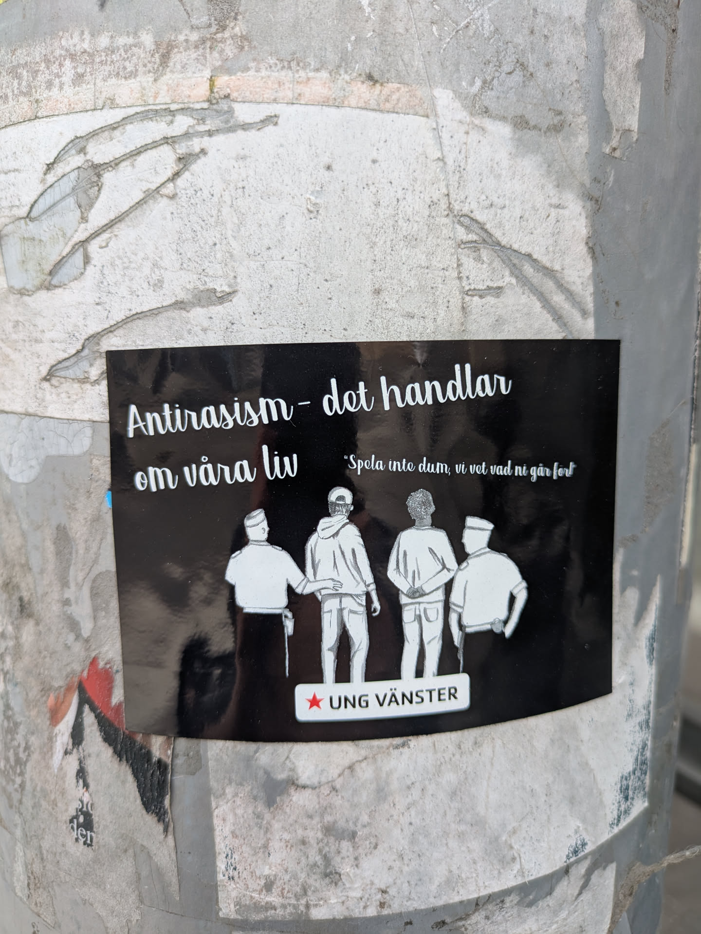 A sticker adhered to a post with the phrase 'Antirasism - det handlar om våra liv' ('Anti-racism - it's about our lives') at the top. Below the text are silhouetted figures of people and the phrase 'Spola inte dum, vi vet vad ni går fort'. The logo of 'UNG VÄNSTER' is at the bottom.