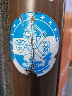 This is a close-up shot of a worn-out sticker affixed to a brown pole. The sticker's main design is a crowned lion, wielding a sword, on a blue and white field, all encircled within a ring of stars.  The sticker shows signs of significant wear and tear; it's cracked and peeling.