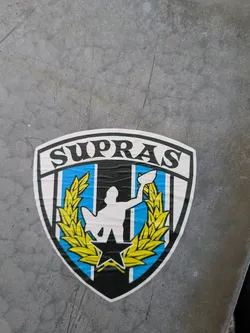 A shield-shaped sticker with the word "SUPRAS" at the top. The shield features a central figure holding a trophy, flanked by wheat, all set against a black, white, and light blue background.  A black star is at the base.