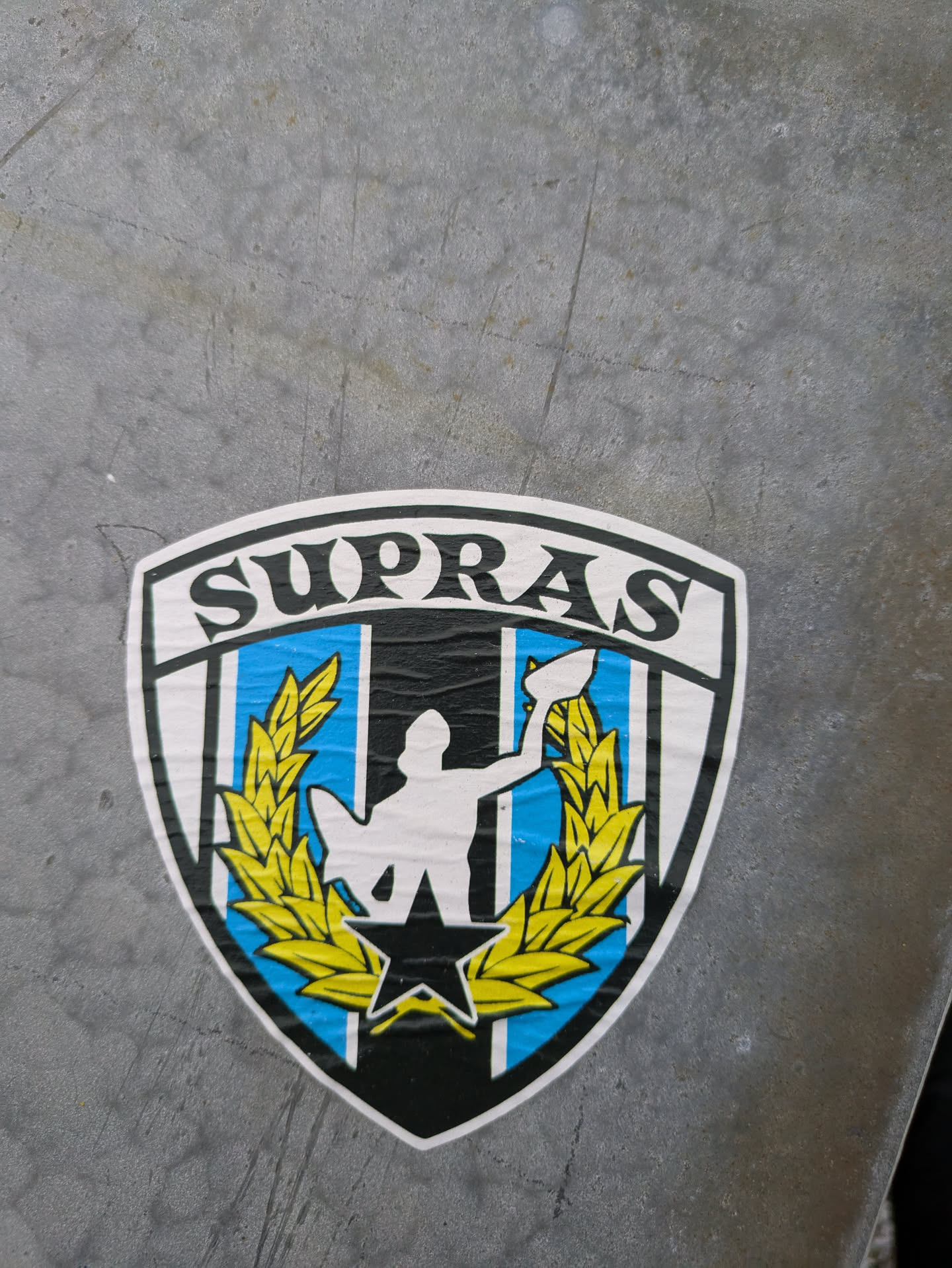 A shield-shaped sticker with the word "SUPRAS" at the top. The shield features a central figure holding a trophy, flanked by wheat, all set against a black, white, and light blue background.  A black star is at the base.