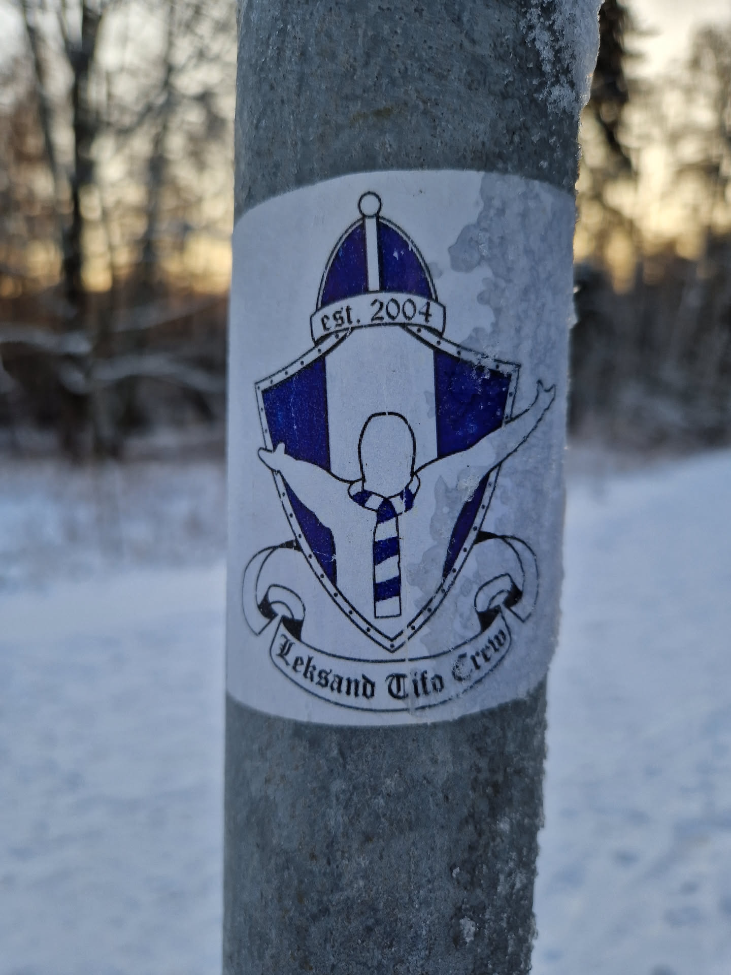 A sticker featuring the Leksand Tifo Crew logo. The logo depicts a person with outstretched arms wearing a striped scarf inside a shield-like emblem. The text 'est. 2004' is visible at the top, and 'Leksand Tifo Crew' is written at the bottom. The sticker appears to be affixed to a metal post covered in frost, suggesting a cold outdoor setting. 