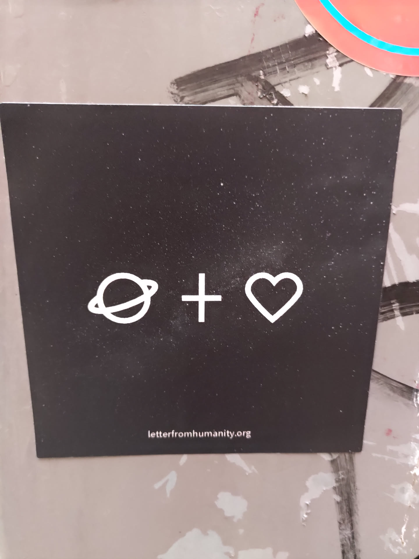 A black square sticker with white minimalist design. It features a planet symbol, a plus sign, and a heart, symbolizing the connection between humanity and Earth. The website letterfromhumanity.org is printed at the bottom.