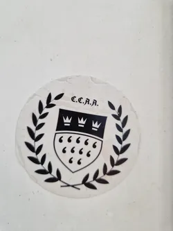A black and white round sticker featuring a crest with three crowns and six quotation marks, encircled by laurel leaves. The acronym C.C.A.A is printed at the top. The sticker appears to be vintage.
