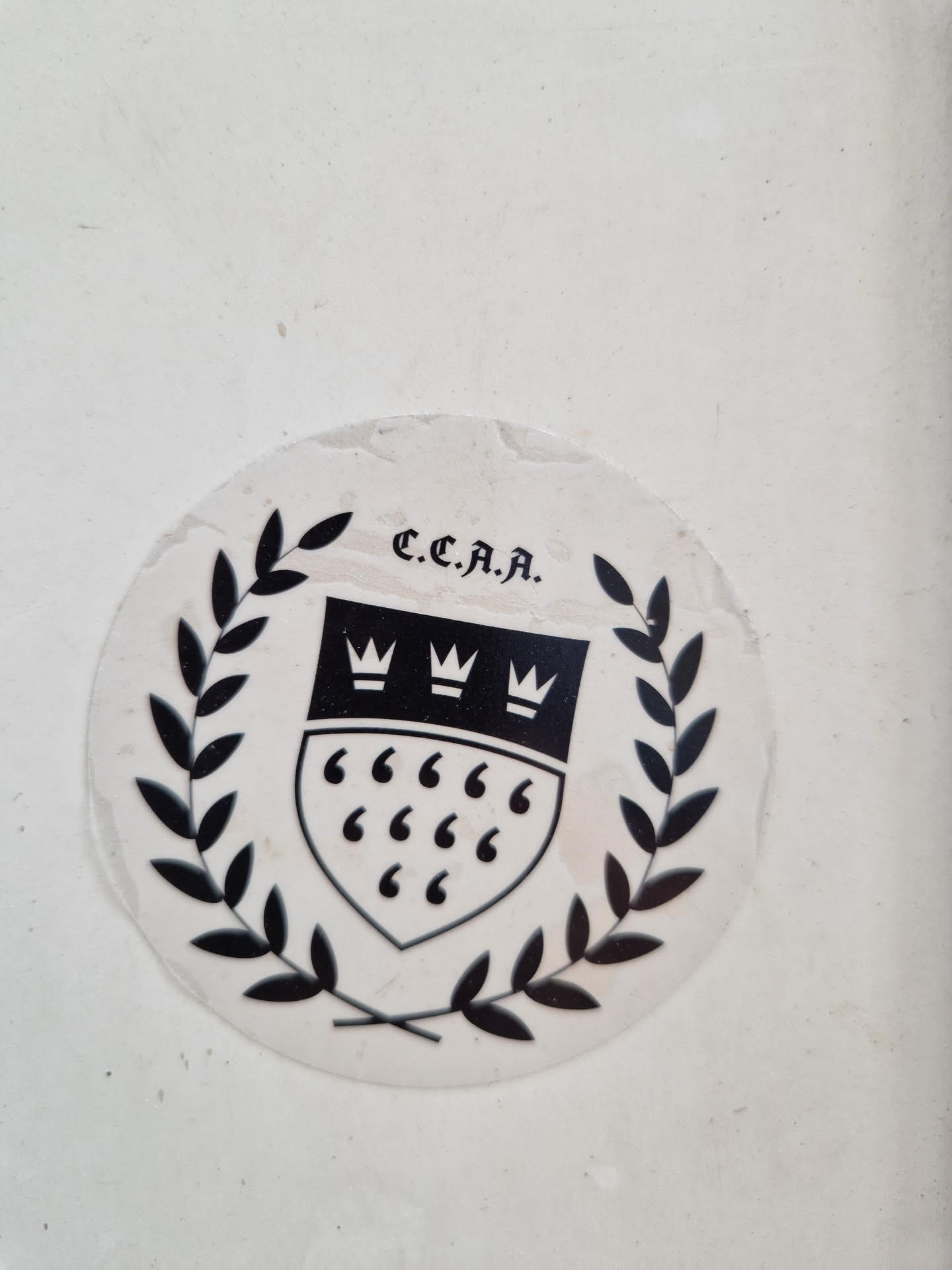 A black and white round sticker featuring a crest with three crowns and six quotation marks, encircled by laurel leaves. The acronym C.C.A.A is printed at the top. The sticker appears to be vintage.