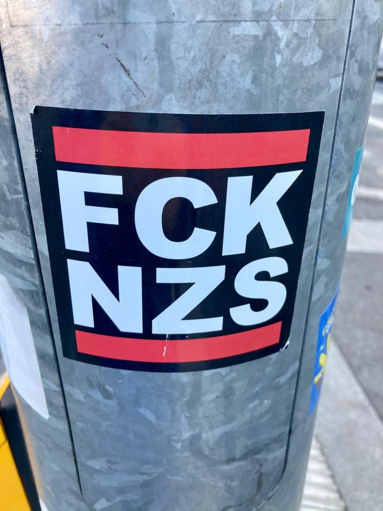 The sticker is rectangular with a black background. It features two prominent horizontal red stripes, one near the top edge and one near the bottom edge. Centered on the black background, between the red stripes, is large, bold white text. The text is arranged in two lines: 'FCK' on the top line and 'NZS' on the bottom line. The sticker is slightly worn and adhered to a grey, metallic pole.