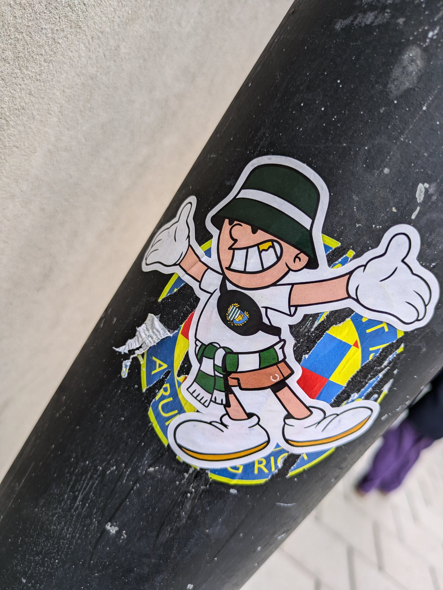 This cheerful sticker shows a cartoon figure with a wide smile, wearing a green bucket hat, a white shirt, and a green and white scarf. The character is giving a thumbs up with both hands. The sticker has some damage on the edges and the lower part is partially covering other stickers that seem to be related to a sports club, given the colours and symbols partially visible.  The background of the main sticker is mostly black, and other visible parts suggest a logo or emblem.