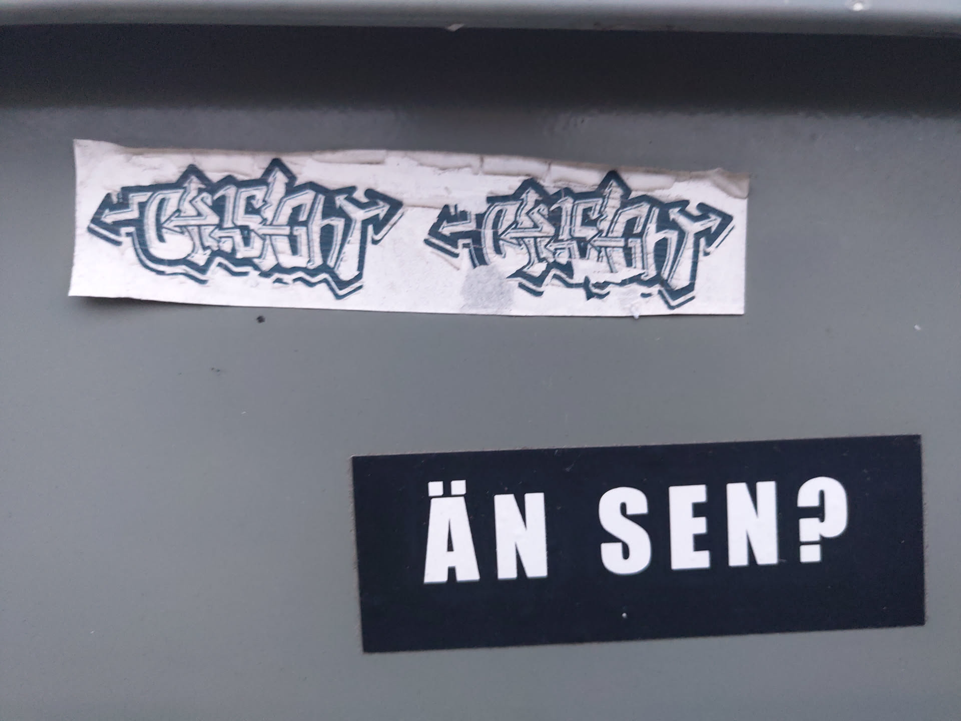 Two stickers are attached to a gray surface. The top sticker features stylized graffiti text that reads "GESICHT" twice, mirrored. The bottom sticker is simpler and displays the phrase "ÄN SEN?" in bold, sans-serif uppercase letters. The stickers appear to be hand-made or independently produced, not mass-produced commercial stickers.